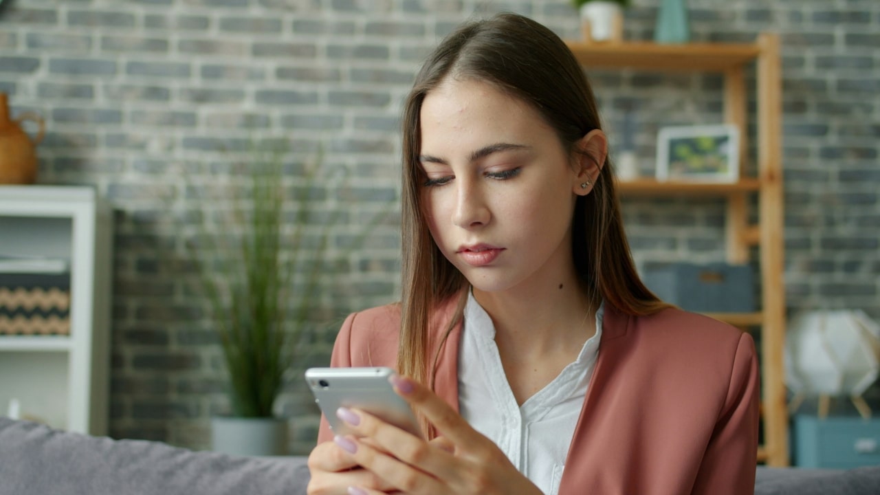 A woman in a pink blazer looks down at her smartphone in a modern room.