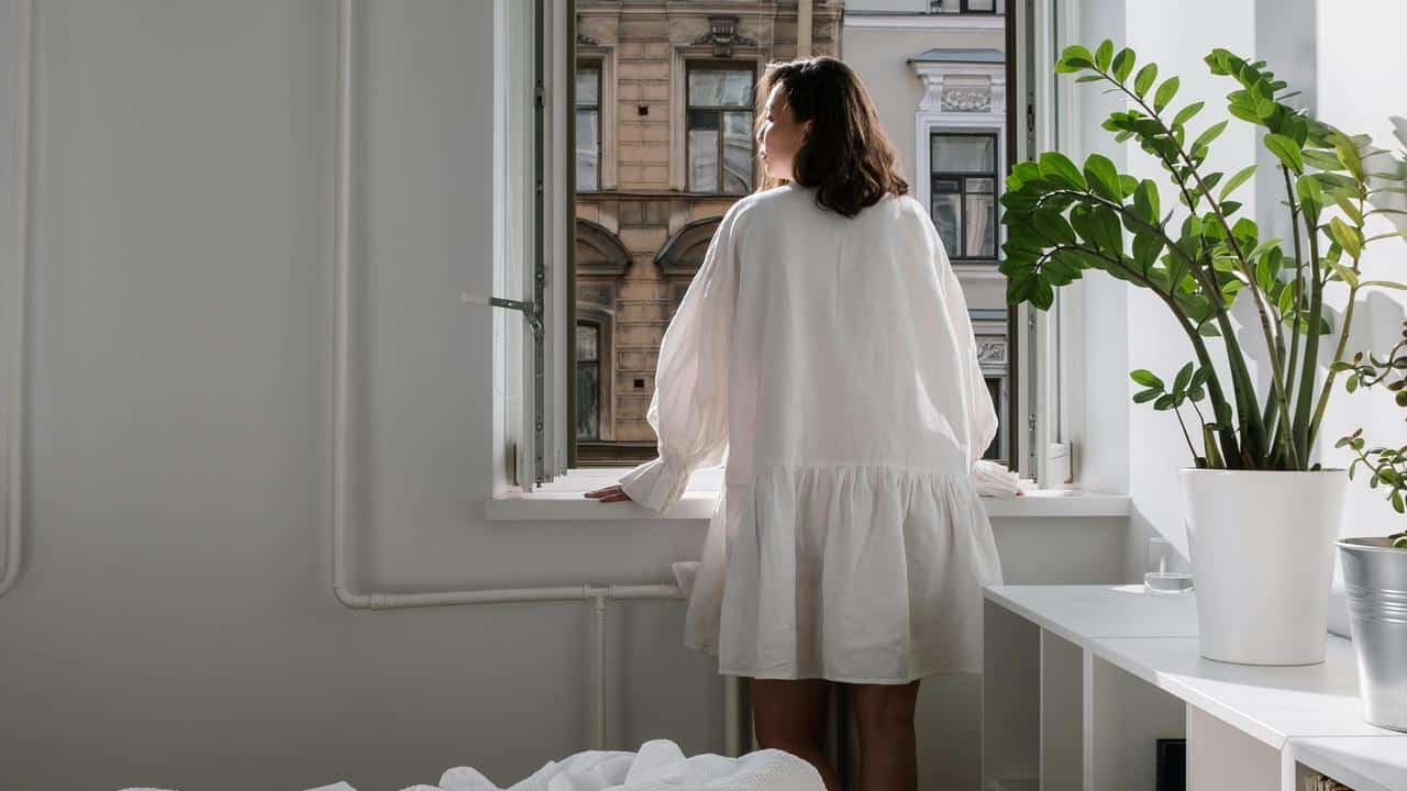 A person in a light dress standing by an open window and looking outside.