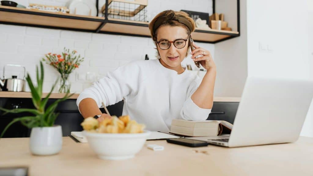 A woman talking on the phone while working on a laptop at home.