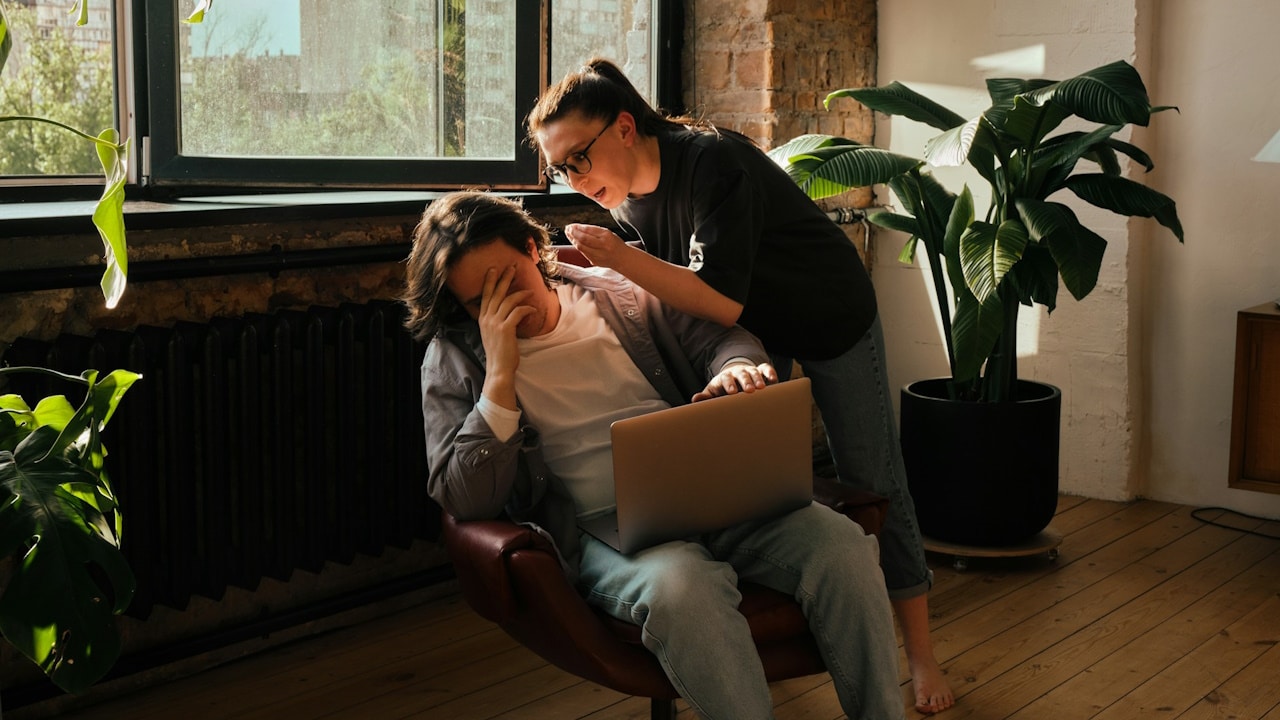 A woman standing and talking angrily to a man sitting with a laptop, who has his face covered with his hand.
