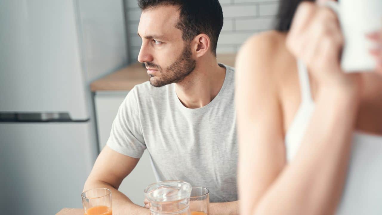 A man looks away in a kitchen, seemingly upset during a conversation with an out-of-focus woman.