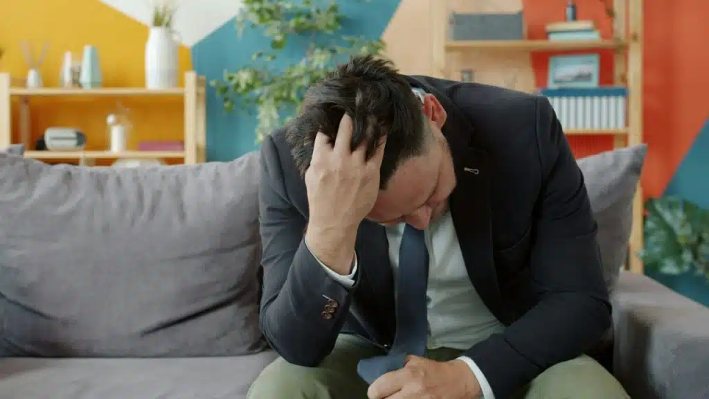 A man in a suit sits on a couch with his head down and hand on hair.