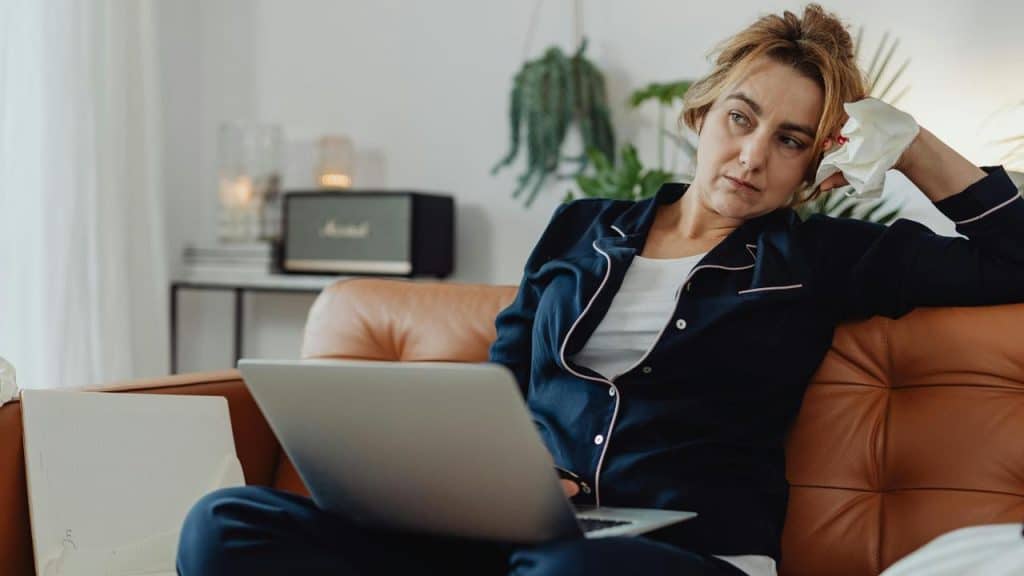 A woman sitting on a couch with a laptop on her lap holding a tissue and looking tired.