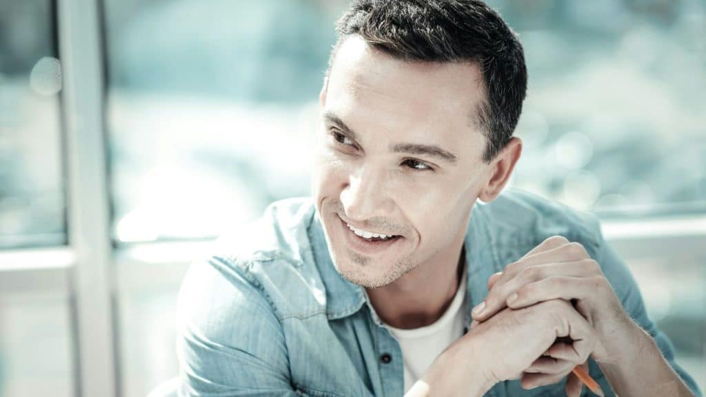 A smiling man in a denim shirt sits with his hands clasped, looking to the side.