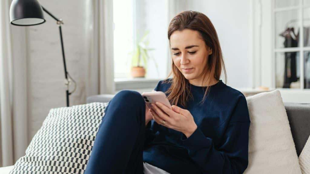 A woman sits on a couch looking at her phone in a bright living room.
