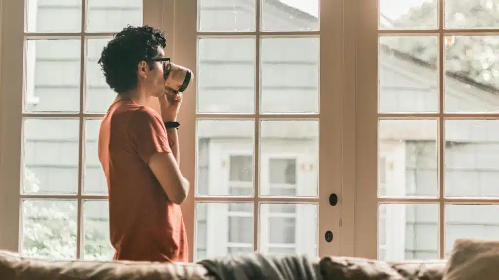 A person stands by a window sipping a drink in daylight.
