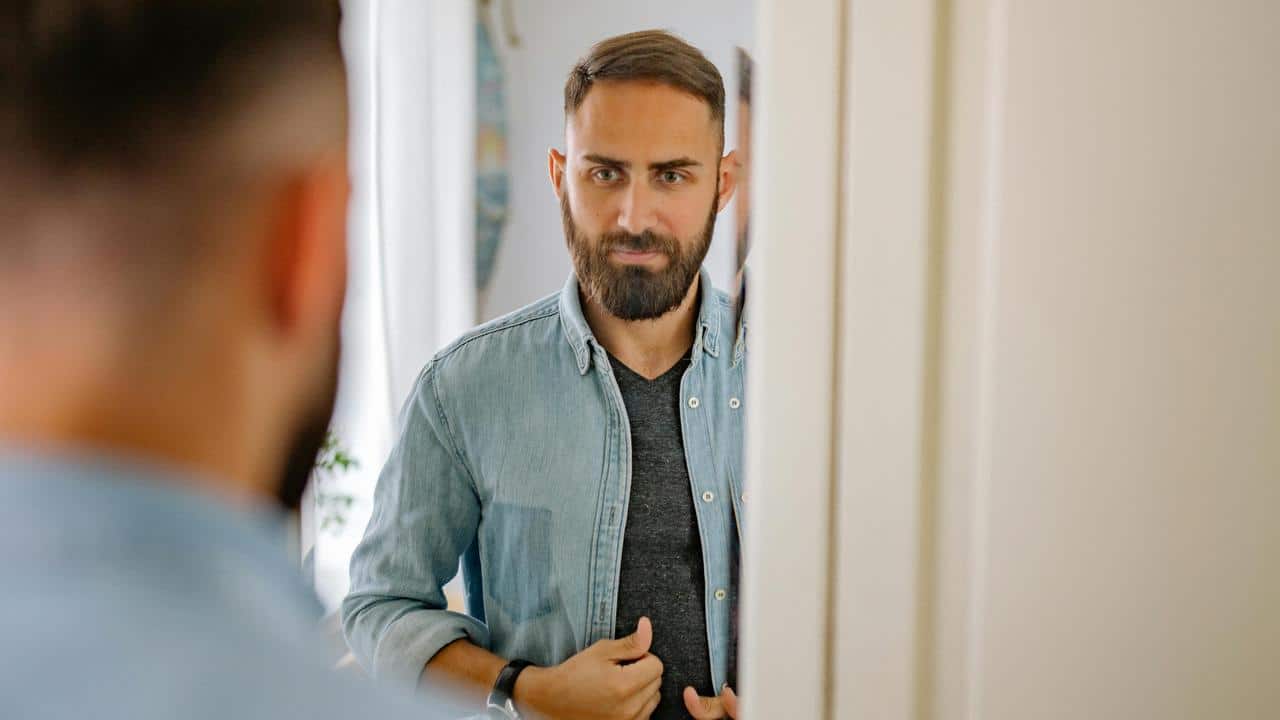A bearded man looking at himself in a mirror indoors.