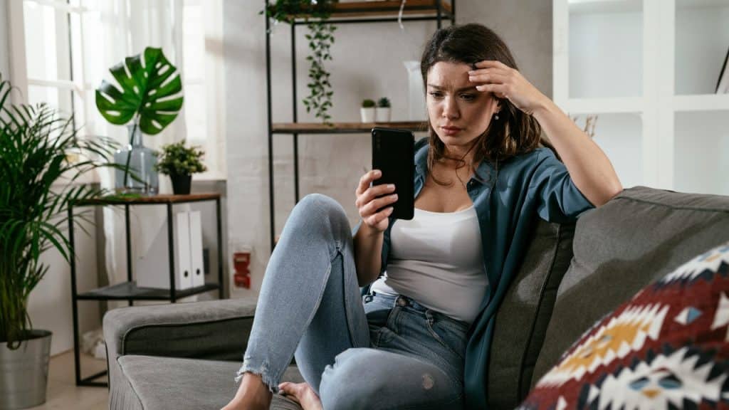 A woman sits on a couch while looking at a smartphone with a concerned facial expression.