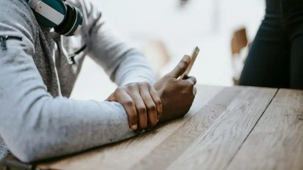 A person sitting at a wooden table holding a smartphone with headphones around their neck.