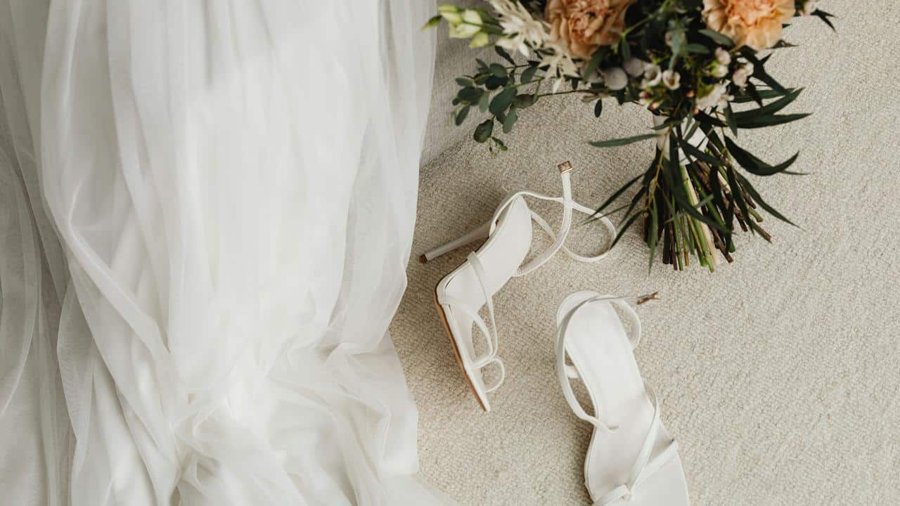 A pair of white heels beside a wedding dress and bouquet.