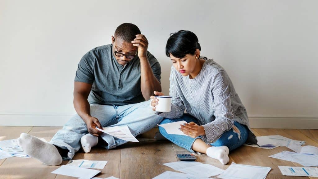 A man and woman sit on the floor surrounded by papers and a calculator.
