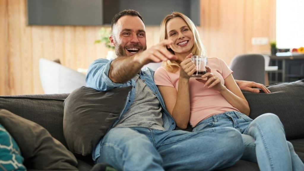 A laughing man and woman sit on a sofa, with the man pointing forward.