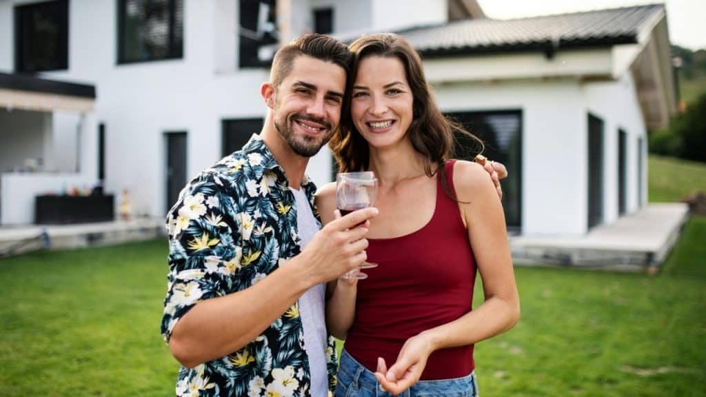 A man and woman smile while holding wine glasses in front of a house.