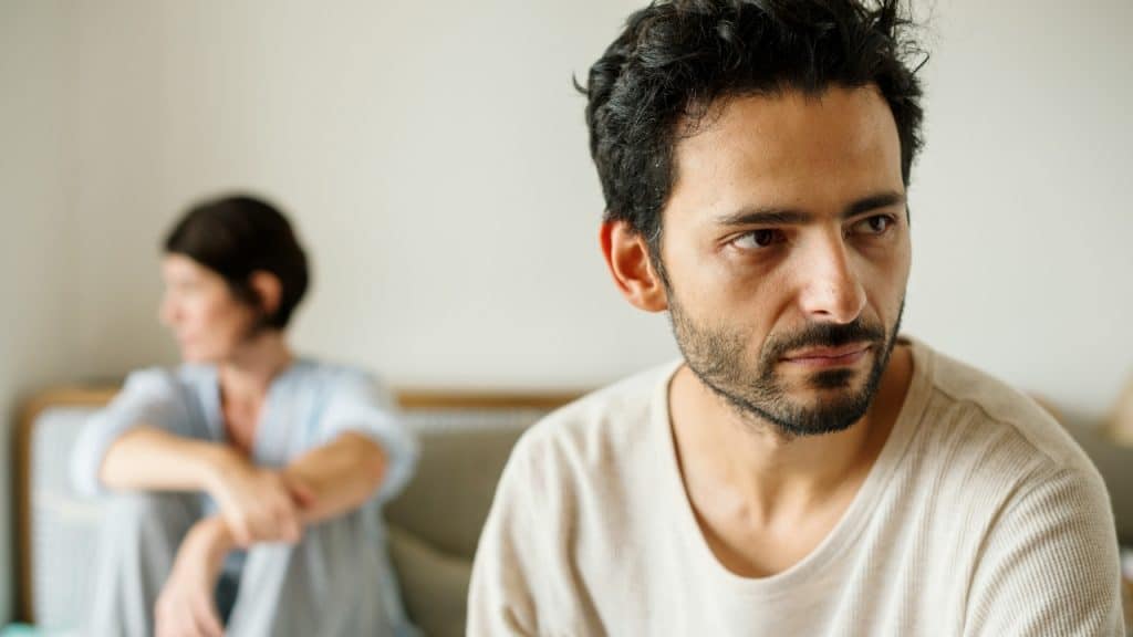 A man with dark hair looks off-camera while a woman sits blurred in the background on a bed.