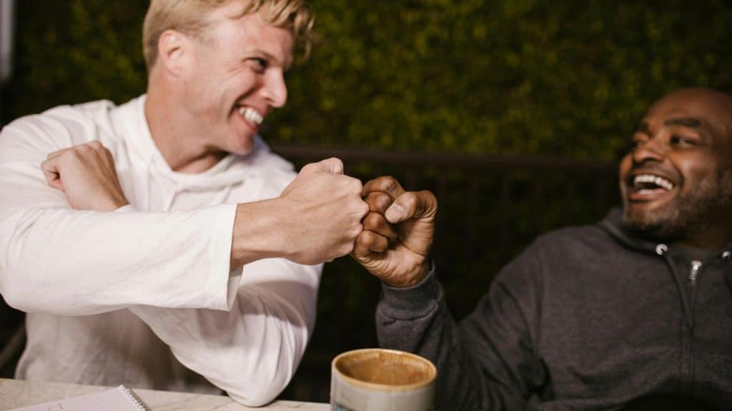 A smiling blond man and another man bumping fists while sitting at a table with a cup nearby.