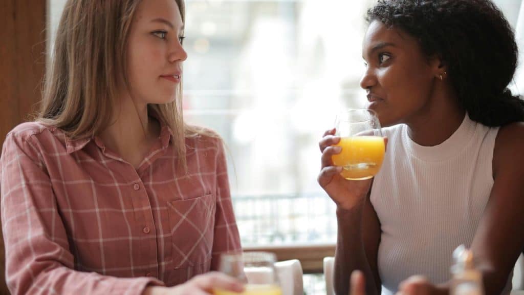A pair of women talking while one drinks a glass of orange juice.
