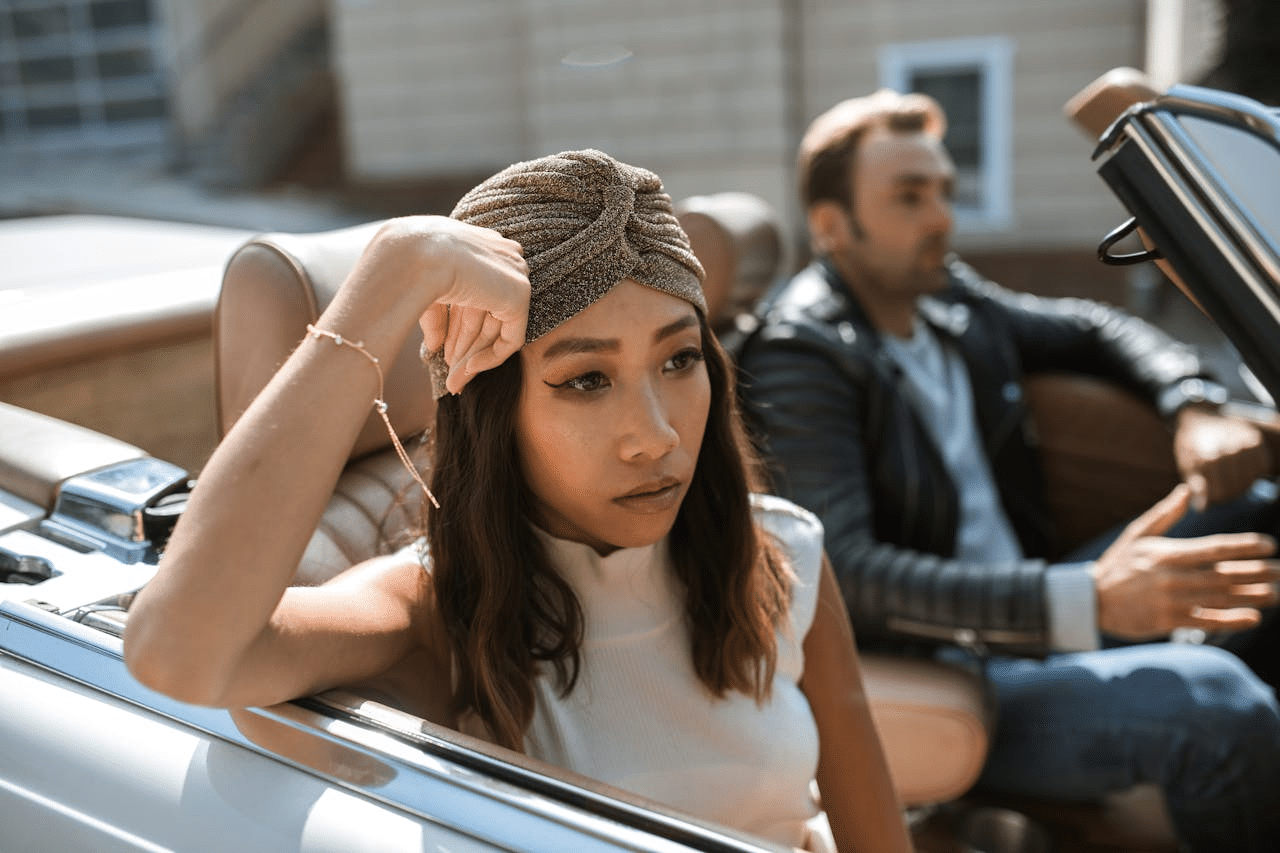 A woman in a white tank top is sitting in a car feeling annoyed while a man is driving the car.