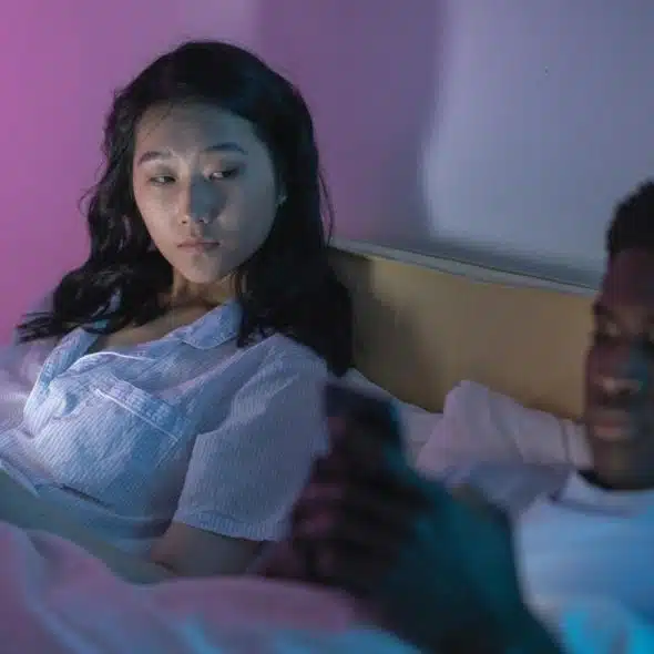 A couple lying in bed at night, both looking at their phones under soft colored lighting.