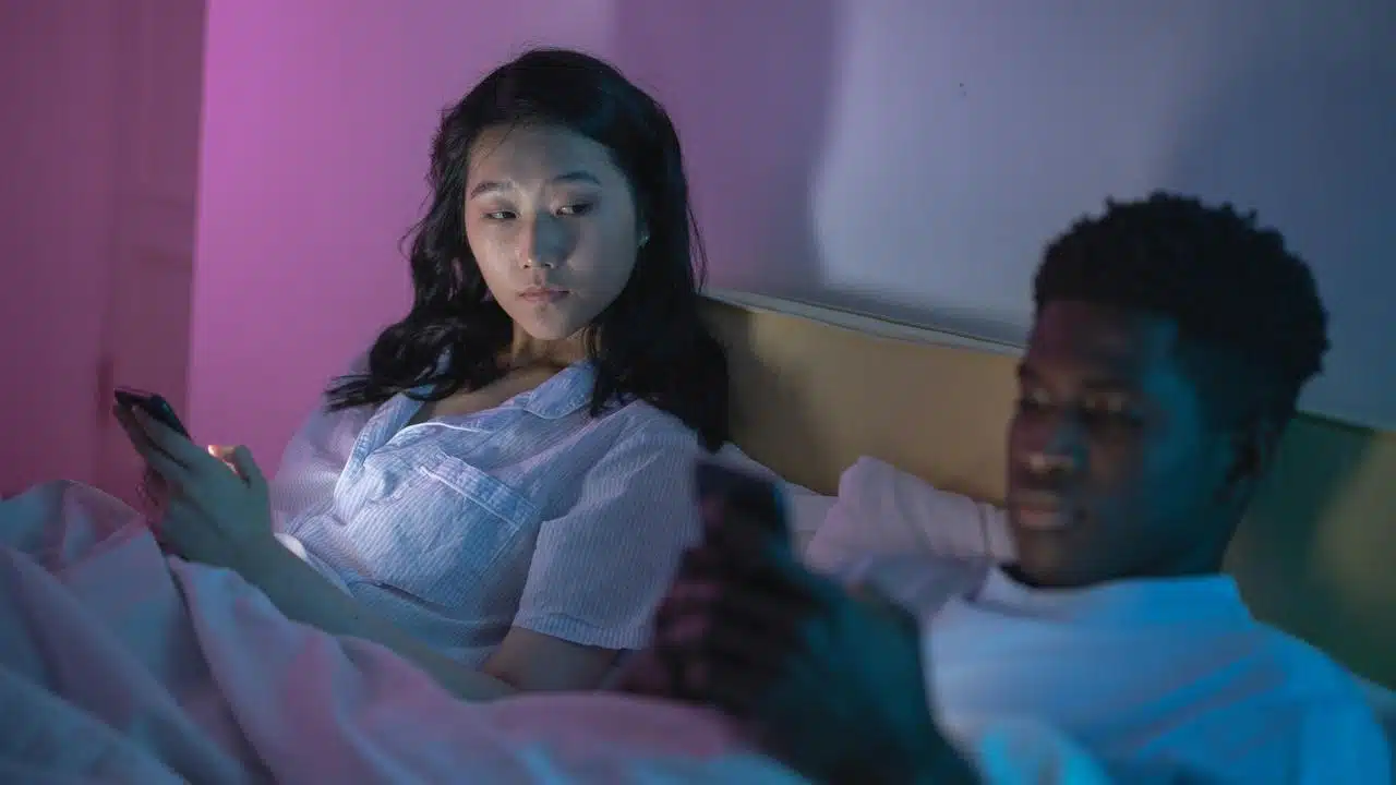 A couple lying in bed at night, both looking at their phones under soft colored lighting.