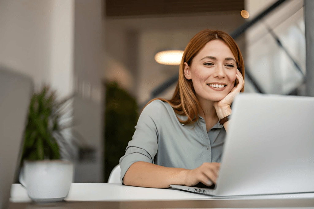 A smiling woman sits at a desk, working on her laptop