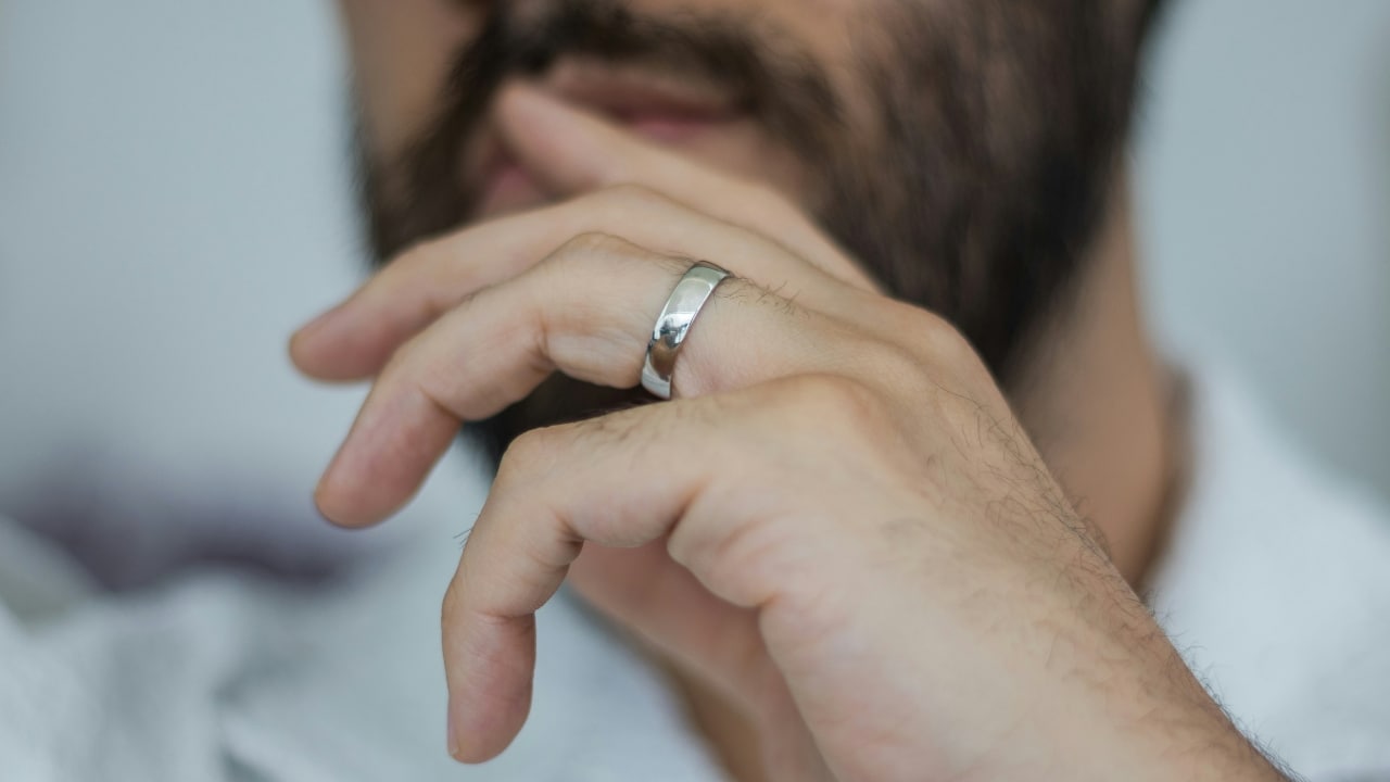 A close-up of a man's hand touching his face, prominently showing a silver band on his finger.