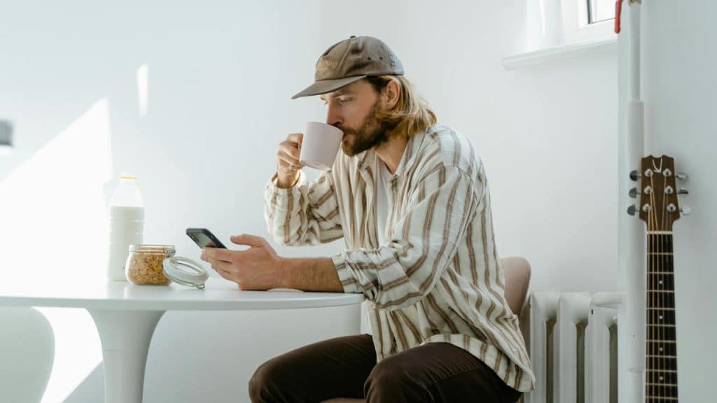 A man drinking from a mug while looking at his phone at a table.
