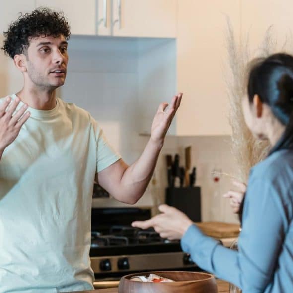 A man with curly hair gestures with both hands while talking to a woman indoors.