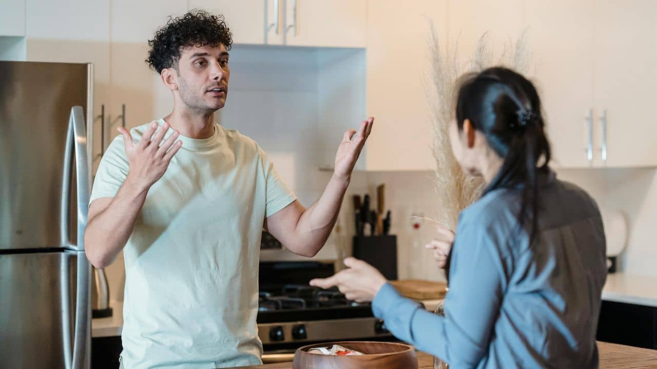 A man with curly hair gestures with both hands while talking to a woman indoors.