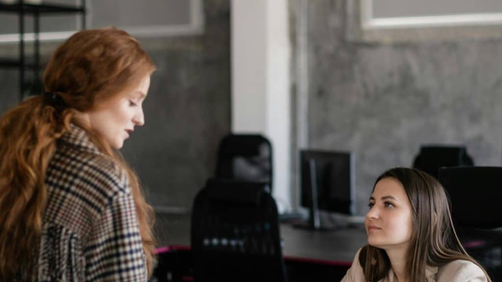 A woman standing and speaking to another seated woman in an office setting.