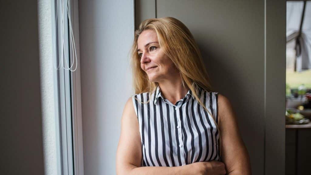 A woman in a striped sleeveless shirt standing by a window and looking outside.