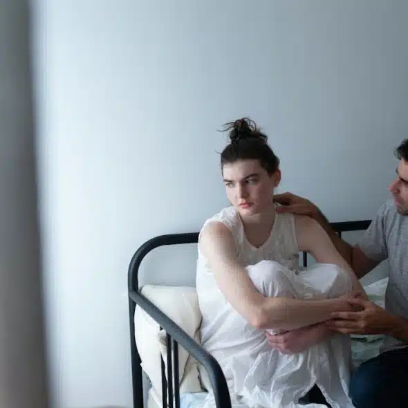 A man gently touches the shoulder of a sad or troubled woman sitting curled up on a metal bed frame.