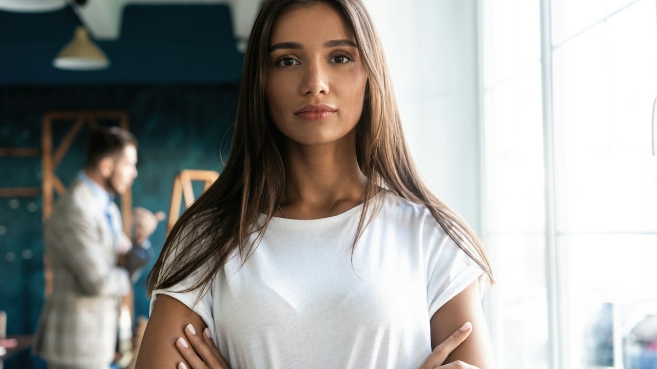 A young woman inside the office is looking at the camera. A man is standing behind her in the background.