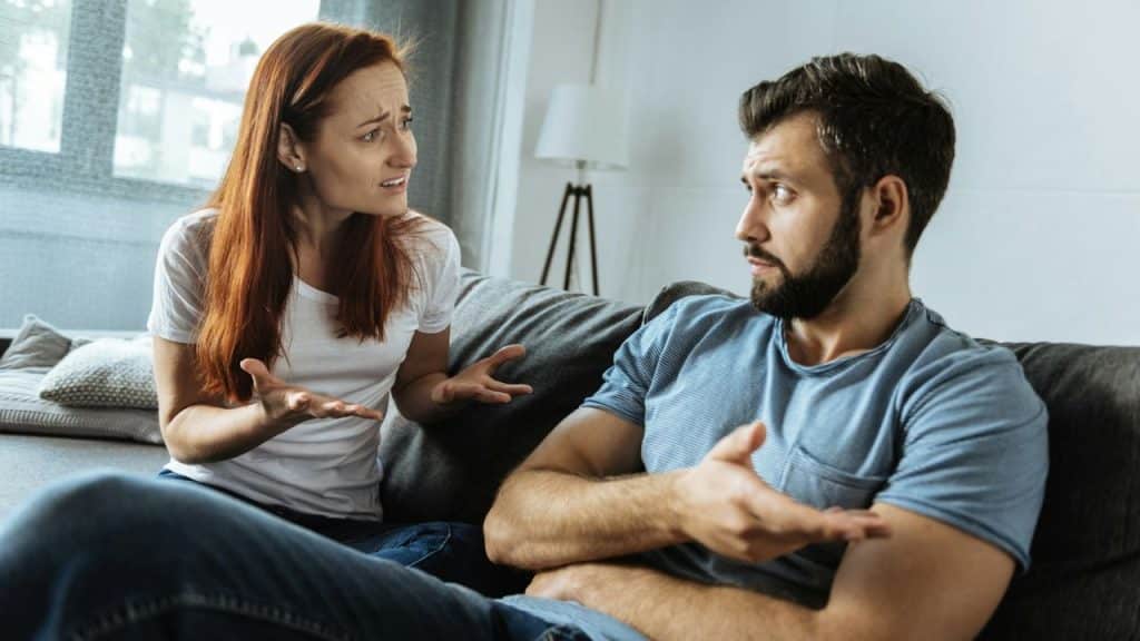 A woman and a man sit on a couch gesturing toward each other with concerned and tense expressions.