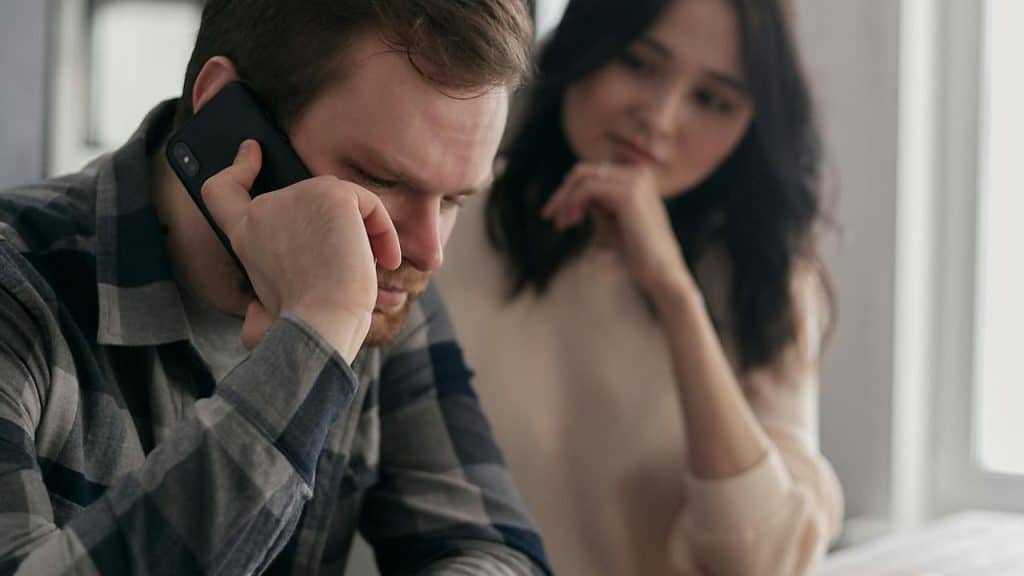 A man talks on a phone while a woman watches nearby indoors.