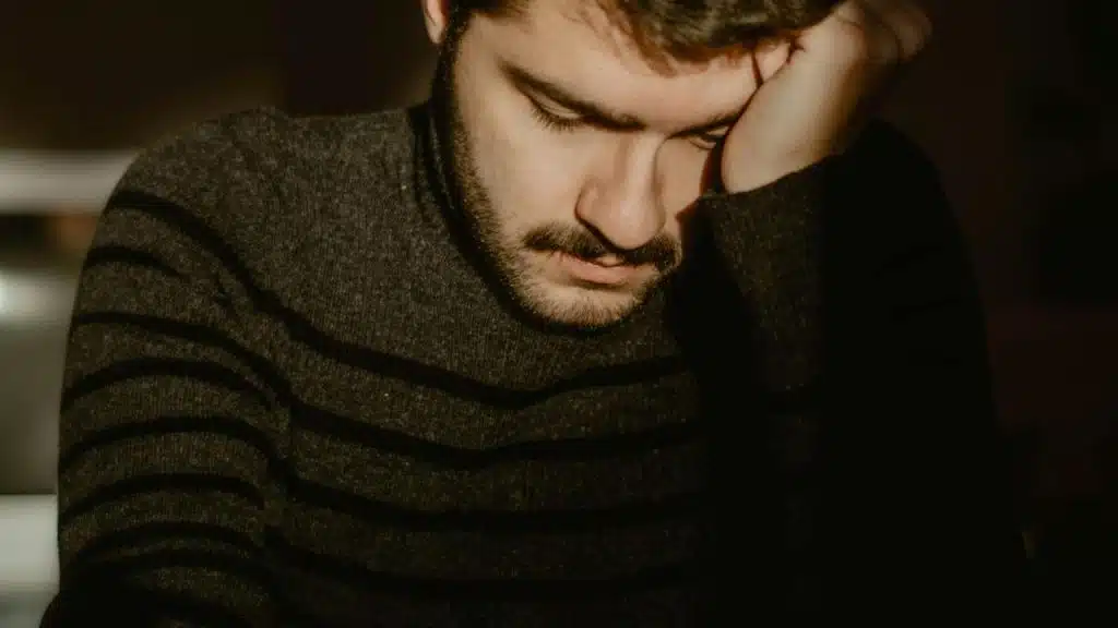 A person rests their head on one hand while looking down in soft light.