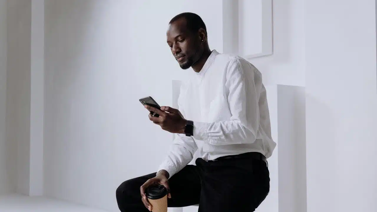 A man in a white shirt looking at his phone while holding a coffee cup.