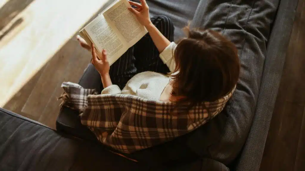 A person sitting on a couch reading a book with a blanket over their shoulders.