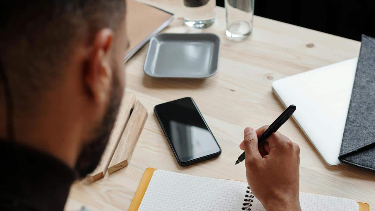 A person writing in a notebook at a desk with a phone and laptop nearby.