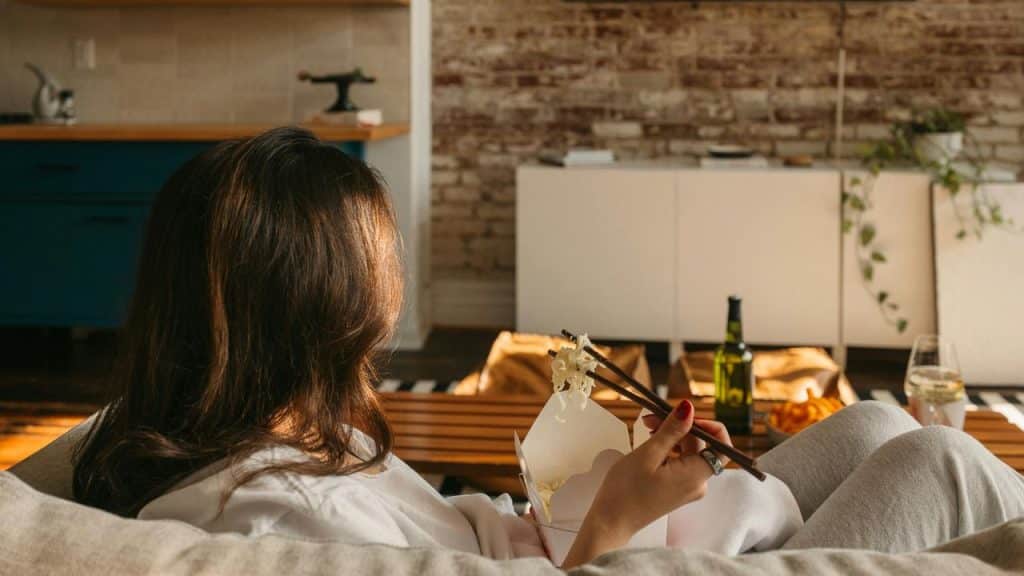 A person relaxing on a couch while eating noodles with chopsticks.