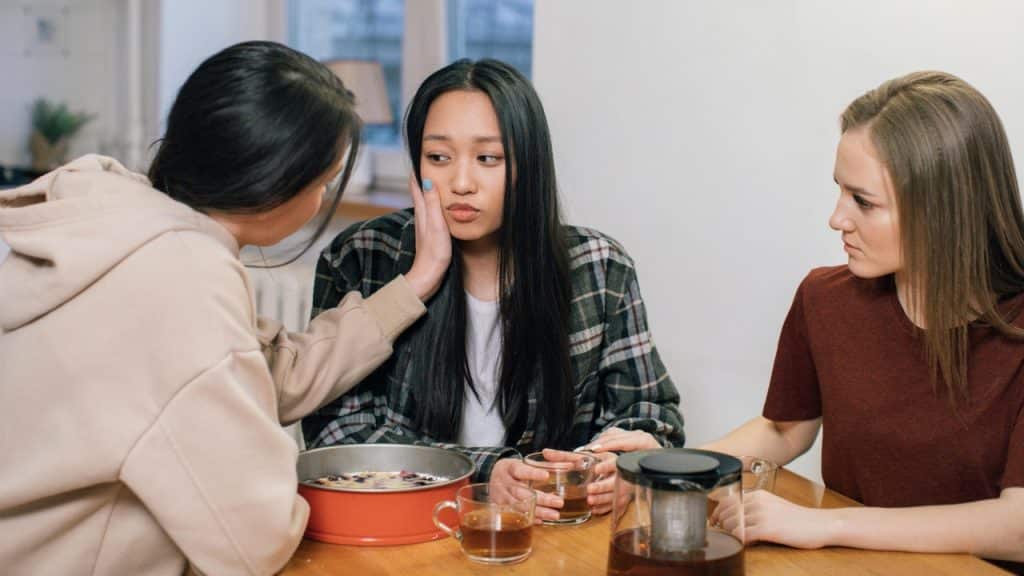 Three women sit at a table with tea; one touches the face of another woman.