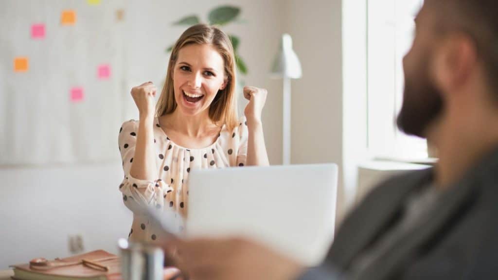 A woman cheers with raised fists while looking at a man behind a laptop.
