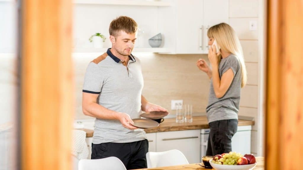 A man carries plates in a kitchen while a woman talks on her phone.
