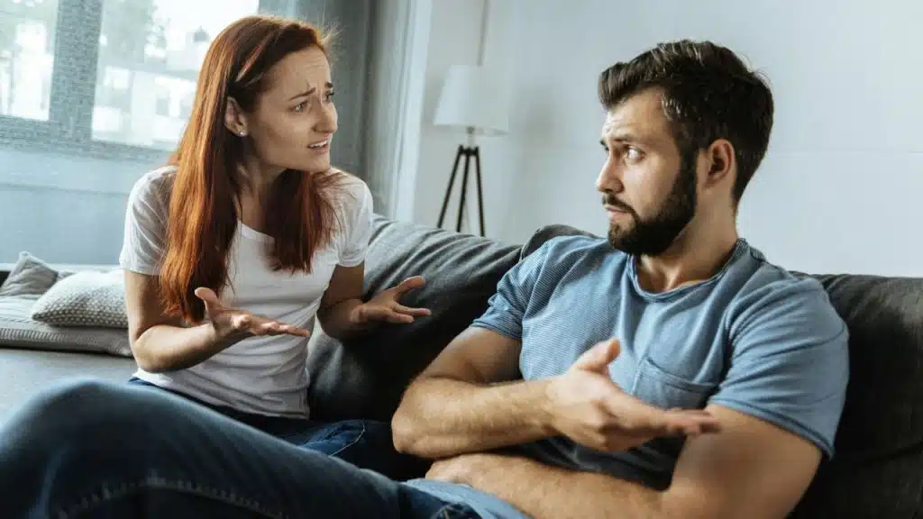 A woman with red hair and a man on a couch gesture while talking.