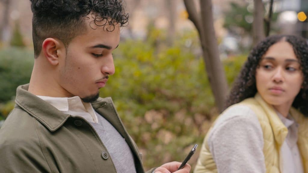 A man looks down at a phone while a woman looks toward him from behind.