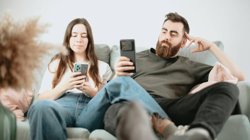 A man and woman sit on a grey sofa while both look at their smartphones.