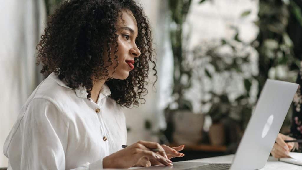 A woman working on a laptop at a desk with plants in the background.
