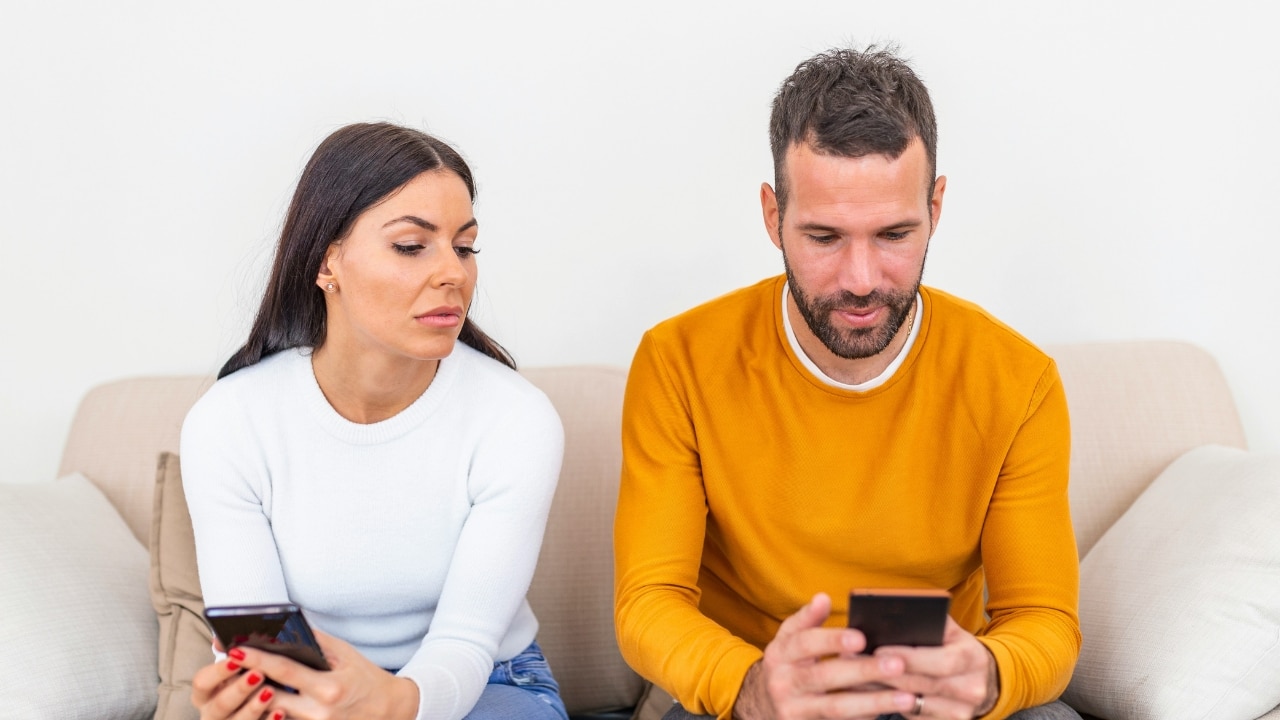 A man and woman sit on a couch, both looking at their own separate smartphones.