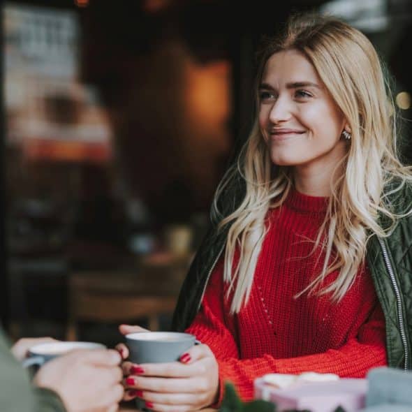 A blonde woman in a red sweater and green jacket smiles while holding a grey mug.