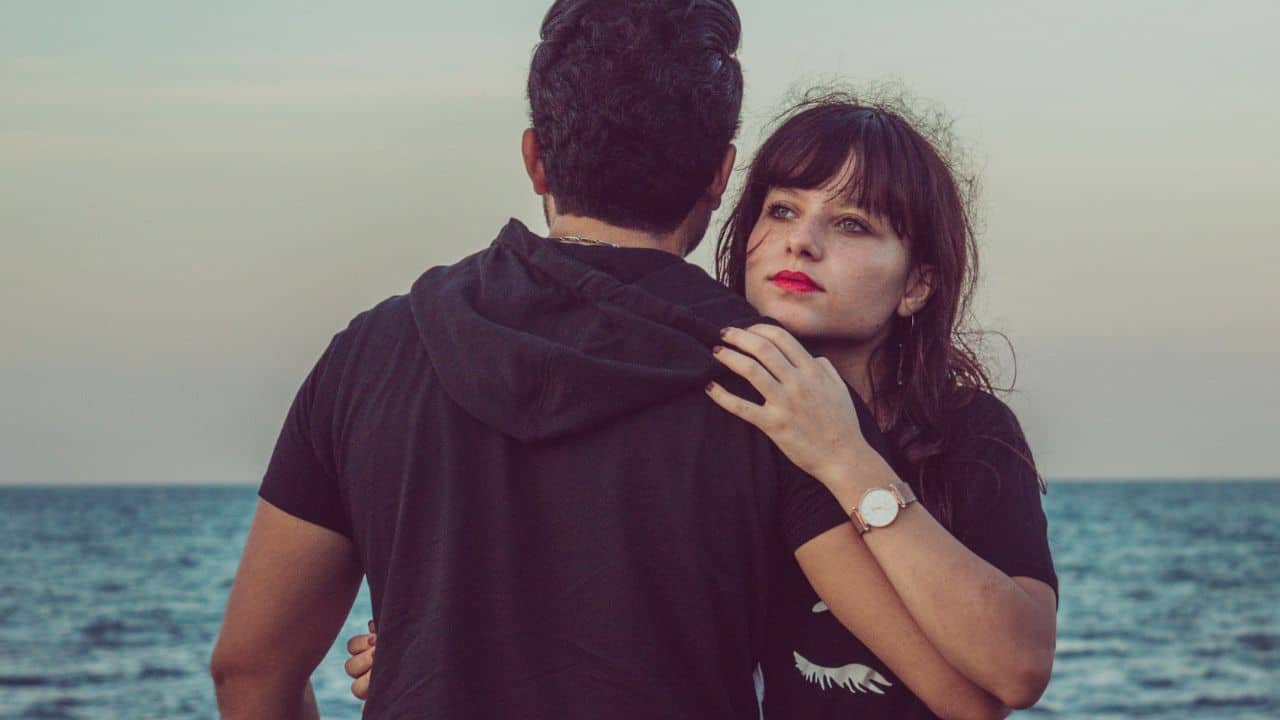 A woman with red lipstick rests her hand on a man's shoulder by the sea.
