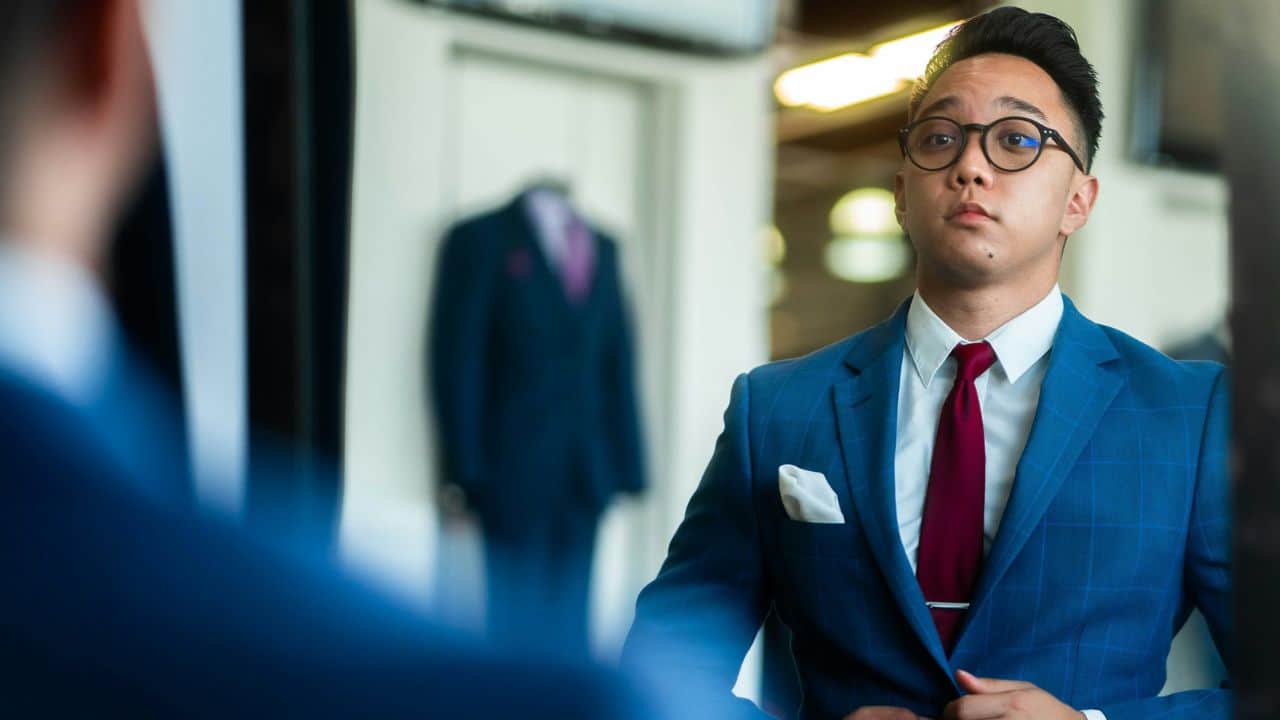 A man in a blue suit and red tie adjusts his clothing while looking in a mirror.